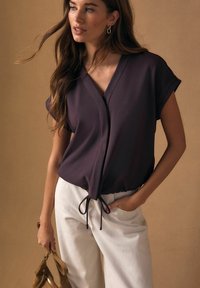 Krátký rukáv, zavinovací top v tmavě švestkové barvě s výstřihem do V a nastavitelným stahovacím lemem. Kombinován s bílými kalhotami. Hladká textura látky.