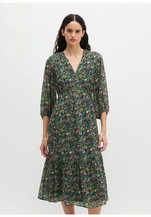 Vrouw met lang donker haar draagt een knielange bloemenjurk met driekwart mouwen en een V-hals tegen een effen lichte achtergrond.