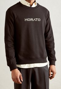Sweat-shirt noir avec poignets et ourlet côtelés, arborant un logo blanc "MORATO" sur le devant. Porté par-dessus une chemise claire avec col.