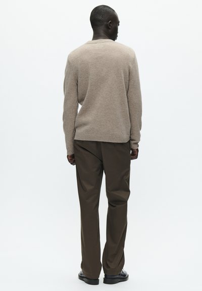 Pull beige en tricot avec un col rond, des poignets et un ourlet côtelés. Associé à un pantalon marron foncé et des chaussures noires.