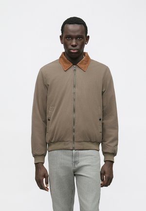 Mees, kes kannab heledat pruuni jakki, millel on kurtšukraed, ja heledaid halli teksaseid, seismas tasase valge tausta ees.