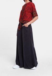 Röd blus med blommönster, korta ärmar och knuten front, matchad med löst sittande svarta byxor. Vita sneakers fulländar outfiten.