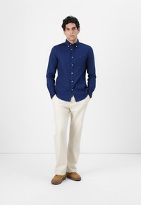 Camisa de botones azul marino de algodón, combinada con pantalones color crema y zapatos marrones. La camisa presenta un cuello puntiagudo y detalles de botones.