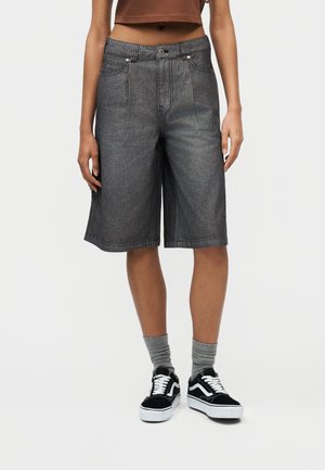 Persona che indossa pantaloncini grigi a righe lunghezza al ginocchio, calzini grigi, scarpe da ginnastica nere e bianche, e un top corto marrone, in piedi contro uno sfondo semplice.