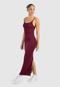 Kobieta w dopasowanej, bezrękawnikowej sukni maxi w kolorze bordowym z bocznym rozcięciem, nosząca białe trampki i skarpetki, stojąca na jasnym tle.