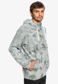 Šedý mikinový pulóver s tie-dye vzorom, so klokankovou vreckom, bielymi šnúrkami a logom na prednej strane. Mäkká textúra a voľný strih.