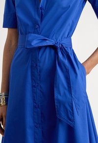 Karališkai mėlyna marškinių suknelė su dirželiu ties juosmeniu, sagomis priekyje, trumpomis rankovėmis ir šoninėmis kišenėmis. Lygi medvilninė medžiaga su struktūrizuota forma.