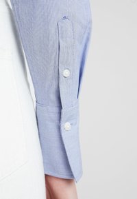 Jasnoniebieska, w paski koszula z długimi rękawami z mankietem, który ma dwa białe guziki, wykonana z teksturowanej tkaniny i z lekko zaokrąglonym brzegiem.