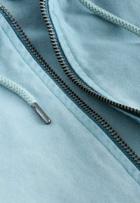 Tissu bleu clair avec une fermeture éclair en métal noir partiellement ouverte et un cordon tressé bleu clair avec une embout métallique.