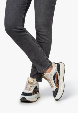 Personne portant un jean slim noir et des baskets blanches avec des détails noirs et bronze, et une doublure en fourrure, croisant les jambes devant un fond blanc.