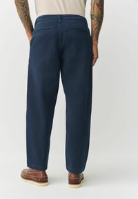 Navy blue, relaxed-fit trousers with side and back pockets, featuring a smooth texture and a casual design. Worn with brown shoes.