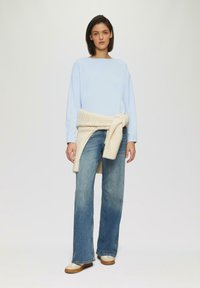 Lichtblauw langemouwen shirt, gecombineerd met lichtblauwe wijde denim jeans en een crèmekleurige gebreide sjaal om de taille. Glad textuur met neutrale kleuren.