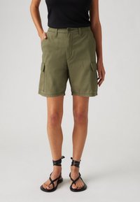 Pantalones cortos cargo verde oliva con un ajuste relajado, cierre de botón frontal, bolsillos laterales y detalles de costura. Combinados con sandalias negras con tiras.