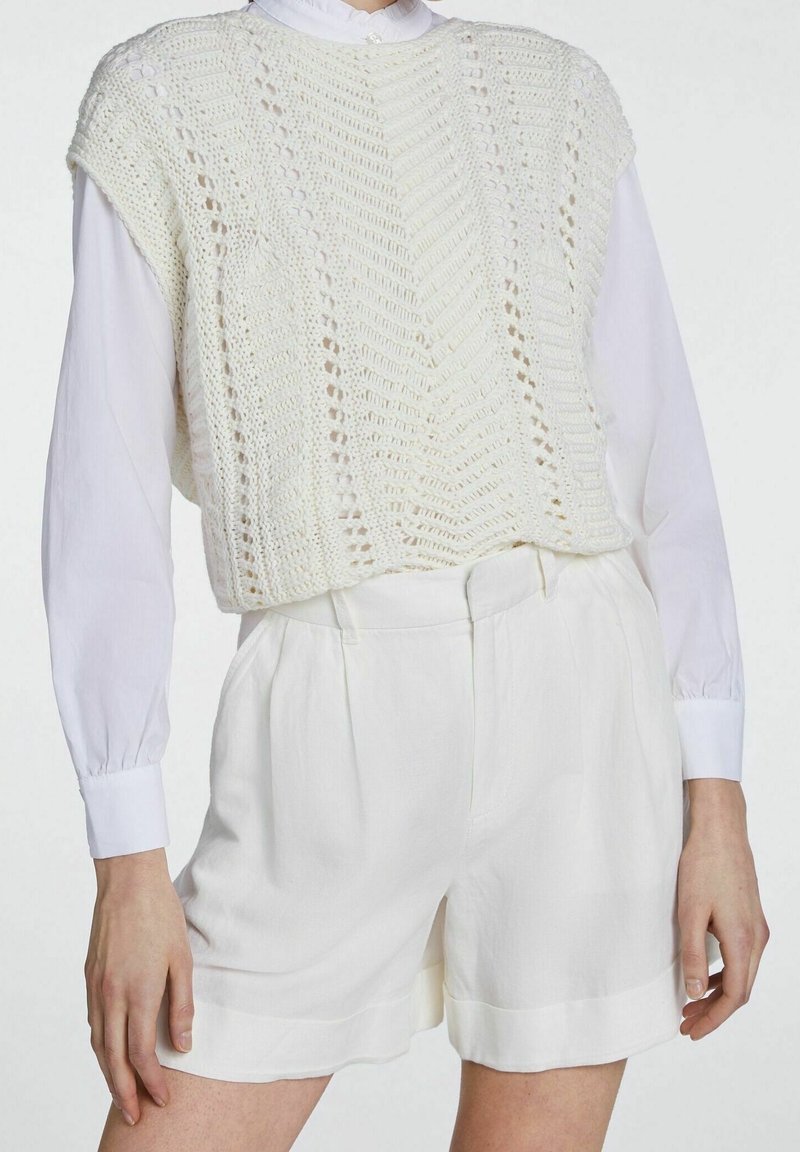 Pull court en tricot blanc avec un motif texturé, superposé sur une chemise blanche à boutons. Associé à un short blanc ajusté.