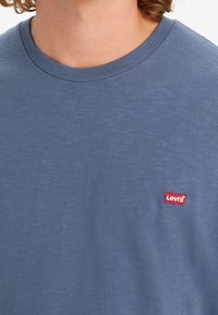 Modrý crewneck tričko vyrobené z jemné bavlny, s malou červenou nášivkou loga Levi's na hrudi, s lehkým a hladkým povrchem.