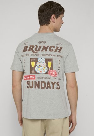 Mann iført grå t-skjorte med brunch-tema grafikk som viser sjømat-tårn, mimoser og reservasjonstider for søndag på baksiden.