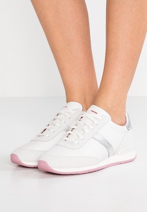 Baskets basses blanches avec semelles roses et accents argentés portées pieds nus, vues de profil et de face.