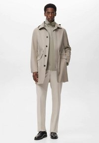 Mand iført beige frakke, lysegrå zip-up sweater, matchende bukser og sorte sko, der står mod en ensfarvet hvid baggrund.