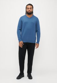 Niebieski sweter z dekoltem w szpic wykonany z gładkiej tkaniny, z długimi rękawami i prążkowanymi mankietami. W połączeniu z czarnymi spodniami i czarnymi sneakersami.