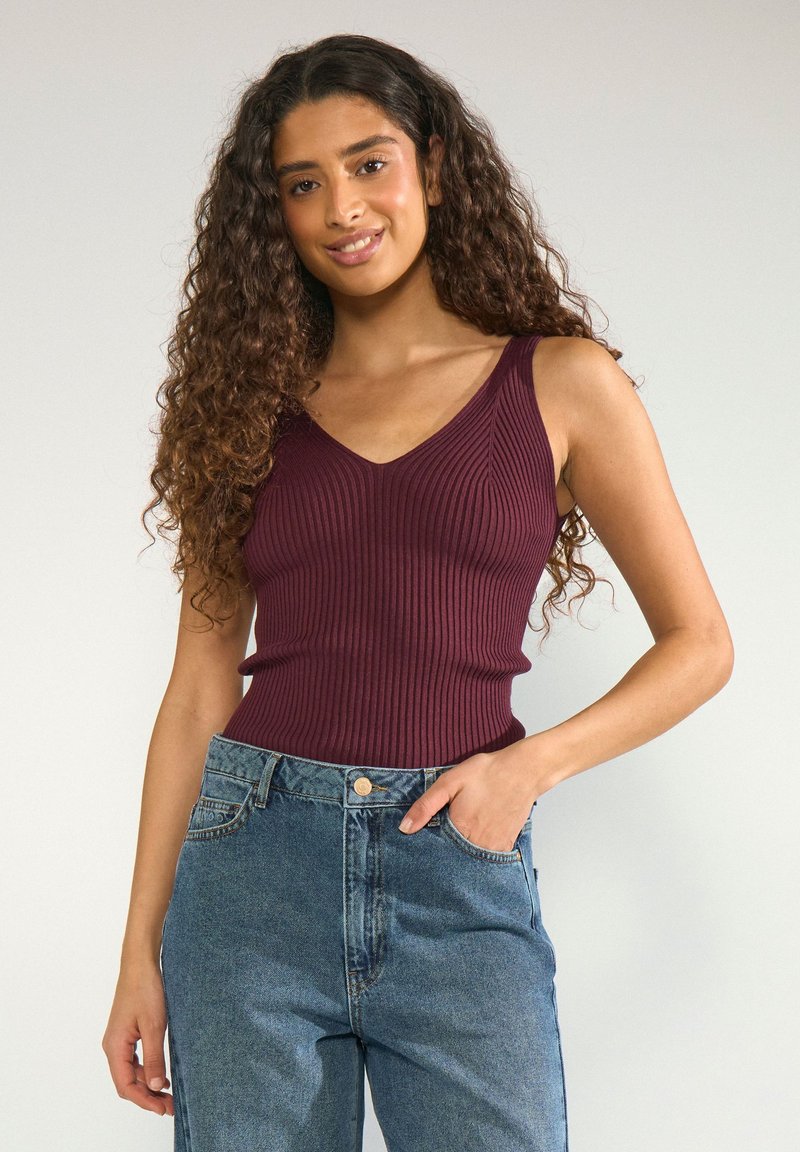 Jeune femme aux longs cheveux bouclés portant un haut sans manches côtelé bordeaux et un jean bleu taille haute, une main dans la poche.