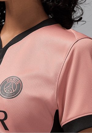 Světle růžový sportovní dres s černými akcenty a výstřihem do V. Má strukturovanou tkaninu a logo s nápisy "PARIS" a "SAINT-GERMAIN".