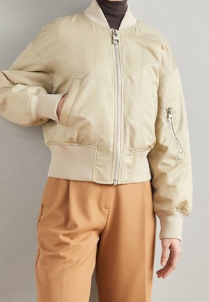 Osoba w kremowej bomberce zapinanej na zamek, z prążkowanymi mankietami i ściągaczem, beżowe spodnie z wysokim stanem oraz ciemny golf pod spodem.