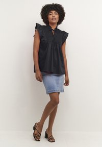 Bluză neagră fără mâneci, cu umeri volonați, detalii smock și guler legat; asortată cu o fustă din denim bleu deschis și sandale negre cu toc pătrat.