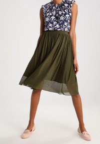 Femme portant un haut sans manches floral bleu marine et une jupe midi olive, assortie à des mocassins rose clair, se tenant contre un fond uni.