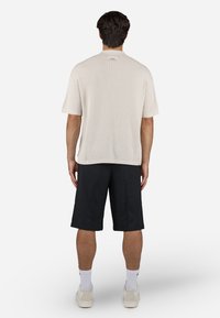 Hombre de pie de espaldas, vistiendo una camiseta de punto de manga corta beige, pantalones cortos negros hasta la rodilla, calcetines blancos y zapatillas beige, sobre un fondo blanco.