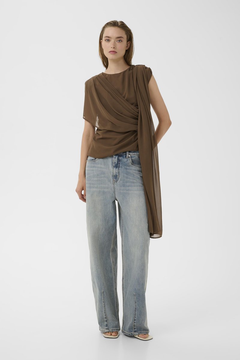 Femme portant un chemisier marron transparent drapé et un jean large bleu clair, debout devant un fond blanc uni.