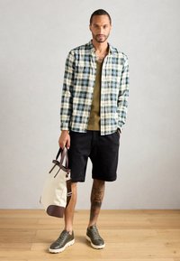 Koszula w kratę w zielono-białe paski, oliwkowy top, czarne szorty, trzymając beżową torbę z brązowymi akcentami, w oliwkowych sneakersach.