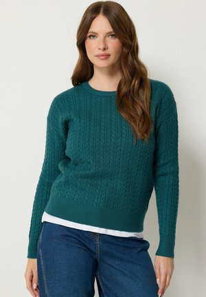 Femme aux longs cheveux bruns portant un pull torsadé bleu sarcelle par-dessus une chemise blanche et un jean bleu, debout devant un fond clair.