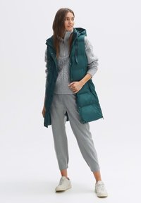 Tassel-Tuch mit Kapuze in Teal, mit Steppstruktur, über einem hellgrauen Strickpullover getragen. Graue, maßgeschneiderte Hose und weiße Turnschuhe runden das Outfit ab.