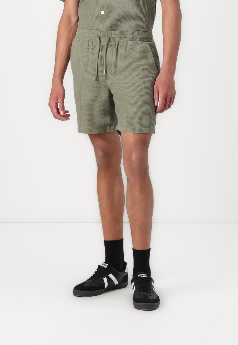Shorts cortos verdes, texturizados, con cintura elasticada y cordón. Combinados con zapatillas negras y calcetines para un look casual.