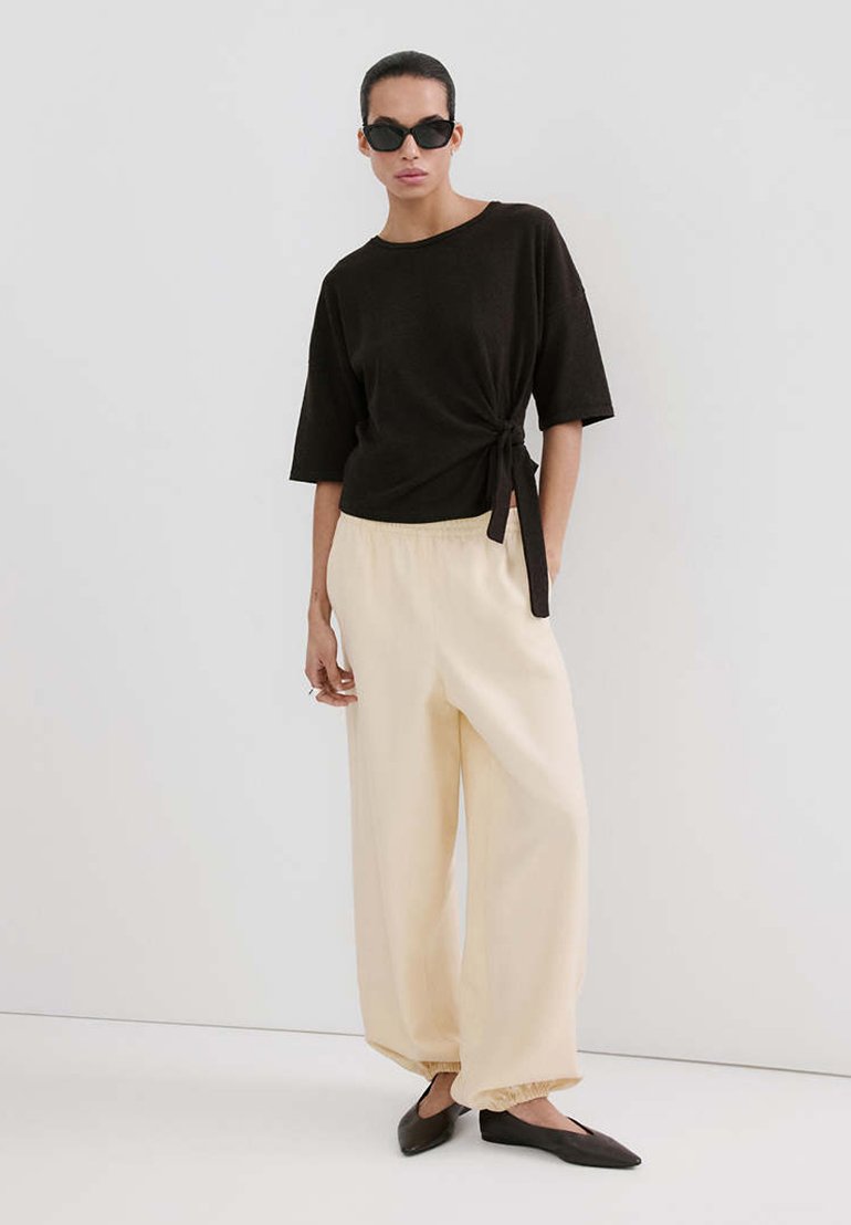 Femme portant un haut noir à manches courtes avec un nouage sur le côté, un pantalon large couleur crème, des ballerines pointues noires et des lunettes de soleil noires, debout devant un fond uni.