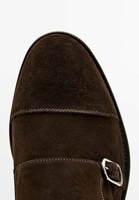 Ruskea mokkanahkakenkä, jossa on pyöreä kärki, koristeellinen soljivarustus ja hillityt tikkauskuviot yläosassa, valkoista taustaa vasten.