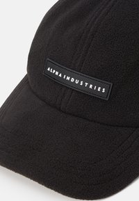 Czarna czapka z polaru z widoczną fakturowaną tkaniną, zakrzywionym daszkiem i prostokątną czarną metką z białym napisem "ALPHA INDUSTRIES".