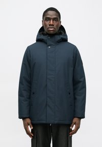Muž v tmavomodrej zimnej bunde s kapucňou a patentnými gombíkmi, stojaci vzpriamene na jednoliatom bielom pozadí.