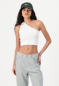 Witte geribbelde crop top met één schouder en een strakke pasvorm, gecombineerd met lichtblauwe en witte gestreepte high-waisted broek. Groene pet voegt contrast toe.
