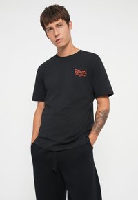Sort bomuld t-shirt med rød "Walt's Waffles" tekstlogo på venstre bryst. Regular fit, korte ærmer, blød tekstur, minimalistisk design.