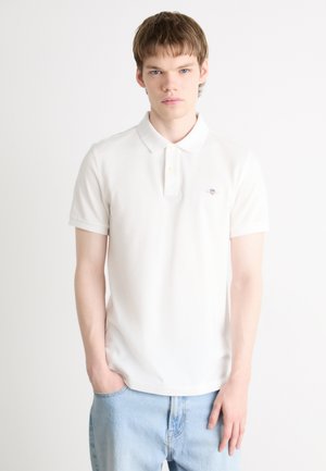 White cotton polo shirt with a three-button placket, short sleeves, and a small logo accent. Paired with light blue denim jeans.