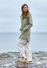 Ljusgrön vattentät jacka med hög krage och framfickor, i kombination med lösa beige linnebyxor och sneakers med gula accenter.