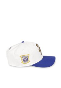 Gorra de béisbol blanca con visera azul, que presenta gráficos bordados en dorado y morado, incluyendo el logo del club en el lado. Material de algodón.