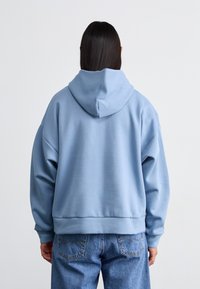 Lysblå hættebluse med afslappet pasform, drop skuldersyninger, ribbede manchetter og bunden af blusen. Glat stoftekstur, minimal detaljer.