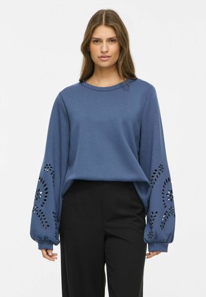 Femme portant un haut bleu à manches longues avec des motifs floraux découpés sur les manches et un pantalon noir, debout devant un fond uni.