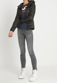 Czarna kurtka puchowa z kapturem, ciemnoniebieska koszula z logo, szare obcisłe dżinsy i białe buty z sznurówkami. Styl minimalistyczny.