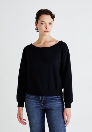 Jeune femme avec des cheveux courts et bouclés portant un pull noir à manches longues et un jean bleu se tenant devant un fond clair.