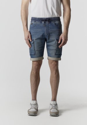 Férfi, kék farmer cargo rövidnadrágban, felhajtott szegéllyel, fehér sportcipőben és fehér pólóban, egyszerű háttér előtt.