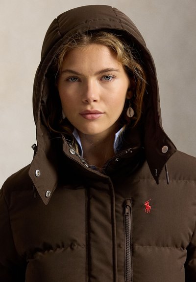 Jeune femme aux cheveux clairs portant une veste d'hiver marron à capuche avec un logo rouge brodé et des boucles d'oreilles en forme de coquillage, fond neutre.