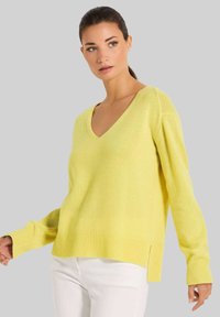 Gelbes V-Ausschnitt-Pullover aus weichem Material mit langen Ärmeln, geripptem Saum und seitlichen Schlitzen. Kombiniert mit weißen Hosen.